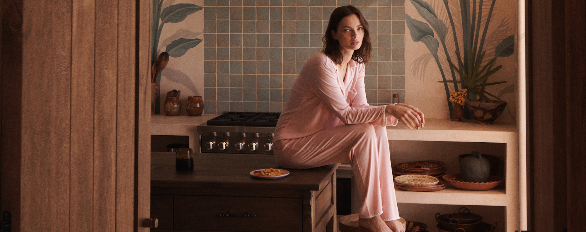 Model wearing Frida Long PJs in Silver Pink/Ivory sitting on counter in kitchen.